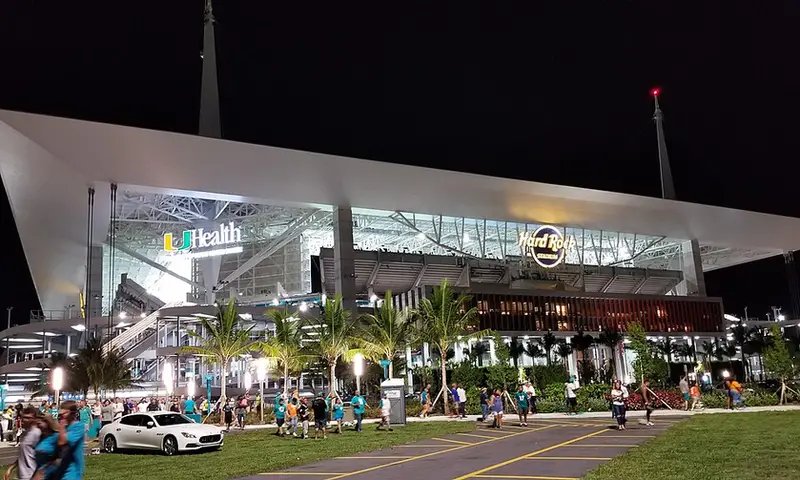 邁阿密花園 Hard Rock Stadium（2026 世界盃 Miami Stadium）封面視覺：夕陽球場外觀與城市背景，前景含公路指標、交通車輛與地圖定位元素，並以圖示呈現賽程、交通、周邊與完整攻略重點
